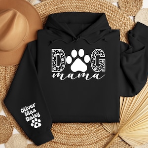 May include: Black hooded sweatshirt with the text "DOG mama" in white with a paw print in the middle of the "O". The letters are decorated with paw prints and bone designs. The left sleeve has the names "Oliver Max Daisy" in white with a paw print below.