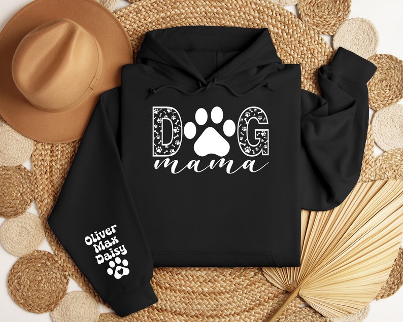 May include: Black hooded sweatshirt with the text "DOG mama" in white with a paw print in the middle of the "O". The letters are decorated with paw prints and bone designs. The left sleeve has the names "Oliver Max Daisy" in white with a paw print below.