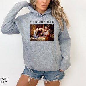 May include: A gray hooded sweatshirt with a pocket on the front. The sweatshirt has a photo printed on the front of a family in Christmas hats sitting around a Christmas tree. The text "YOUR PHOTO HERE" is printed above the photo.