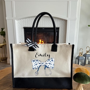 May include: A white canvas tote bag with black trim and handles. The bag features a blue and white floral bow with a stethoscope and the name "Emily" printed in black.
