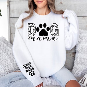 May include: White sweatshirt with black text that reads "DOG mama" with a paw print in the middle of the word "DOG". The letters are filled with a black and white paw print pattern. The left sleeve has black text that reads "Oliver Max Daisy" with a paw print below the text.