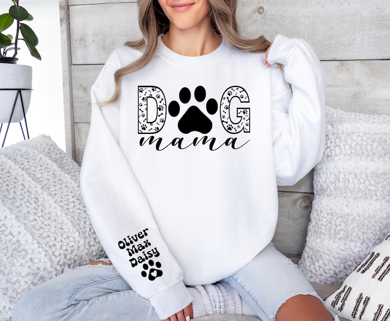 May include: White sweatshirt with black text that reads "DOG mama" with a paw print in the middle of the word "DOG". The letters are filled with a black and white paw print pattern. The left sleeve has black text that reads "Oliver Max Daisy" with a paw print below the text.