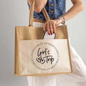A woman is holding a large beige tote bag with the text "Girl's Trip" printed on it. She is also holding a cell phone in her hand.