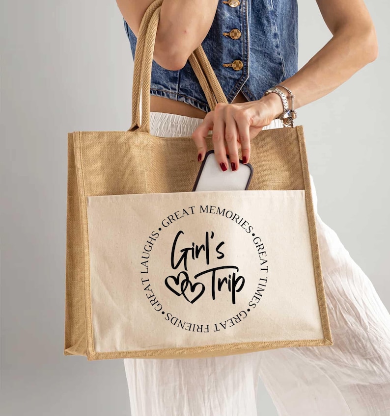 A woman is holding a large beige tote bag with the text "Girl's Trip" printed on it. She is also holding a cell phone in her hand.