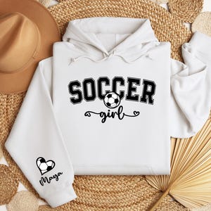 May include: White hoodie with black text that says "Soccer girl" and a soccer ball graphic. The left sleeve has a black heart with a soccer ball graphic and the name "Maya" written below it.