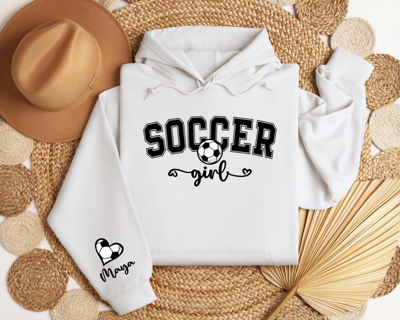 May include: White hoodie with black text that says "Soccer girl" and a soccer ball graphic. The left sleeve has a black heart with a soccer ball graphic and the name "Maya" written below it.