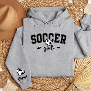 May include: Gray hooded sweatshirt with the text "Soccer girl" and a soccer ball graphic. The sleeve has a heart with a soccer ball and the name "Maya" written below.