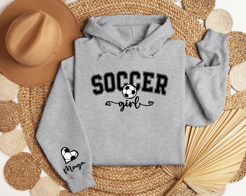 May include: Gray hooded sweatshirt with the text "Soccer girl" and a soccer ball graphic. The sleeve has a heart with a soccer ball and the name "Maya" written below.