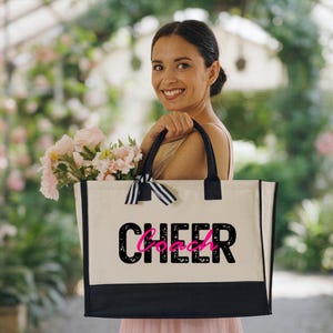May include: A canvas tote bag with black handles and a black bottom panel. The word "CHEER" is printed in black, with the word "Coach" in pink, on the front of the bag. A black and white striped bow is tied to the handle.