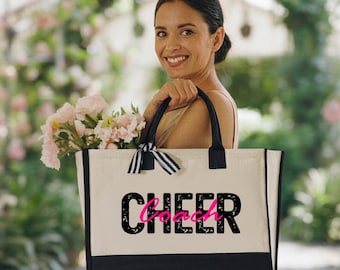 May include: A canvas tote bag with black handles and a black bottom panel. The word "CHEER" is printed in black, with the word "Coach" in pink, on the front of the bag. A black and white striped bow is tied to the handle.