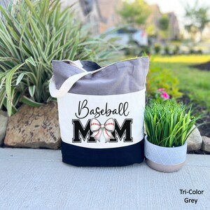 May include: A grey and white canvas tote bag with black handles. The bag has a white panel with the text "Baseball Mom" in black lettering and a white baseball with red stitching tied in a bow. The bottom of the bag is black.