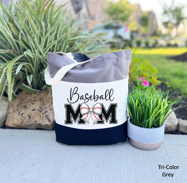 May include: A grey and white canvas tote bag with black handles. The bag has a white panel with the text "Baseball Mom" in black lettering and a white baseball with red stitching tied in a bow. The bottom of the bag is black.