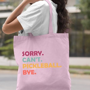 May include: Light pink tote bag with the text "SORRY. CAN'T. PICKLEBALL. BYE." in colorful letters. The bag has a simple design and is carried over the shoulder.