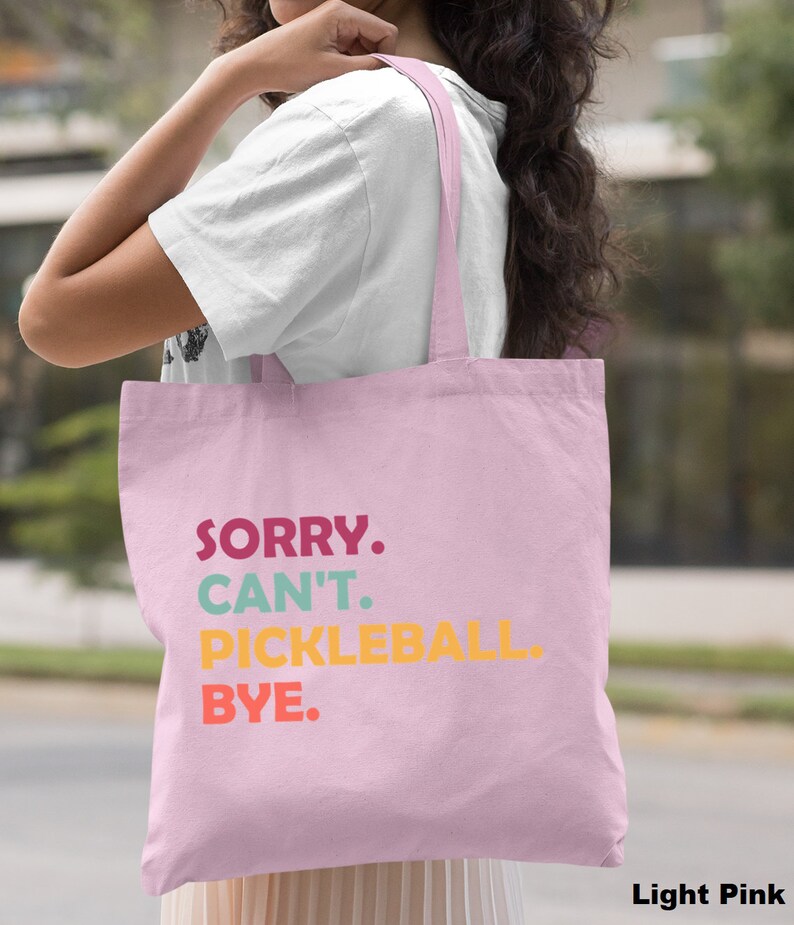 May include: Light pink tote bag with the text "SORRY. CAN'T. PICKLEBALL. BYE." in colorful letters. The bag has a simple design and is carried over the shoulder.