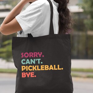 May include: Black tote bag with the text "SORRY. CAN'T. PICKLEBALL. BYE." in colorful letters. The bag is carried over a person's shoulder. The word "Black" is printed at the bottom right corner of the bag.