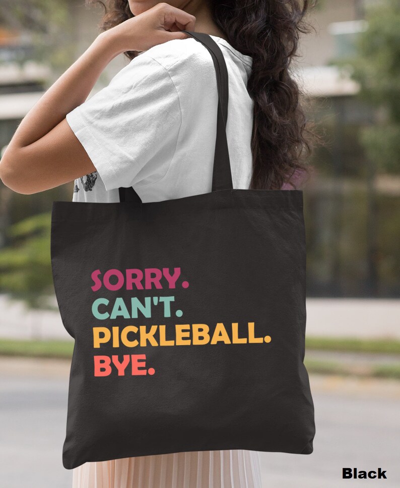May include: Black tote bag with the text "SORRY. CAN'T. PICKLEBALL. BYE." in colorful letters. The bag is carried over a person's shoulder. The word "Black" is printed at the bottom right corner of the bag.