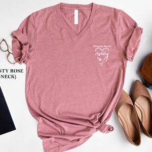 Pu&ograve; includere: Maglietta a collo a V color rosa polvere con la scritta "Hospice Nurse" e un cuore con uno stetoscopio al suo interno. Il nome "Ashley" &egrave; scritto all'interno del cuore.