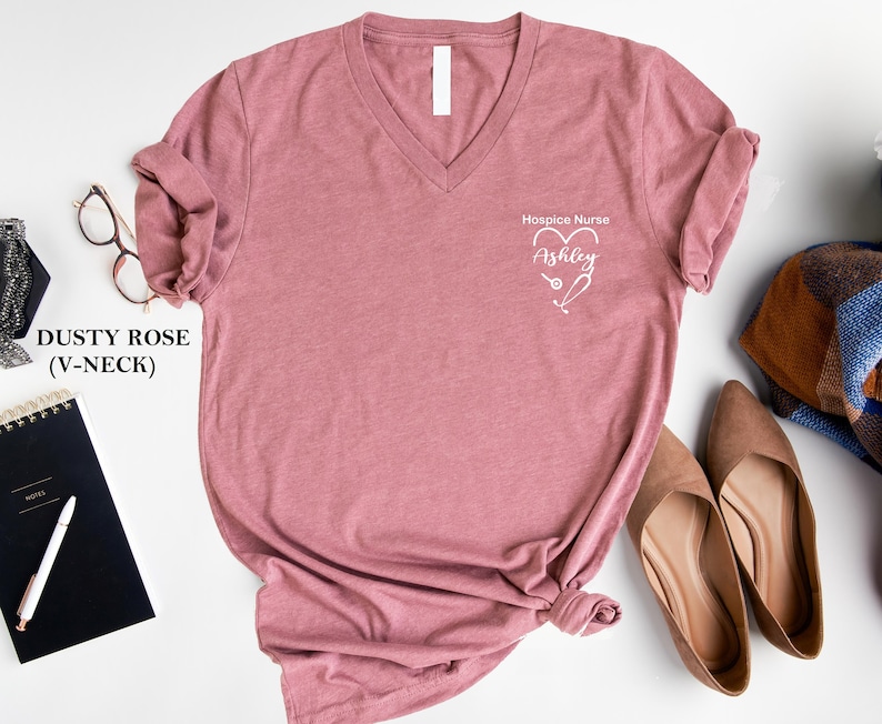 Pu&ograve; includere: Maglietta a collo a V color rosa polvere con la scritta "Hospice Nurse" e un cuore con uno stetoscopio al suo interno. Il nome "Ashley" &egrave; scritto all'interno del cuore.