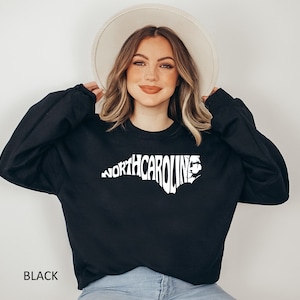 K&ouml;nnte beinhalten: Ein schwarzer Pullover mit einem wei&szlig;en Aufdruck des Bundesstaates North Carolina, wobei das Wort "NORTH CAROLINA" in Form des Bundesstaates ausgeschrieben ist.