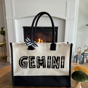 May include: A black and white canvas tote bag with black handles and a black and white striped bow. The bag has the word "GEMINI" printed on the front in black with white stars and other decorative elements.