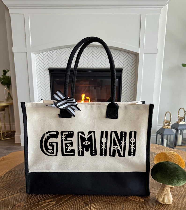 May include: A black and white canvas tote bag with black handles and a black and white striped bow. The bag has the word "GEMINI" printed on the front in black with white stars and other decorative elements.
