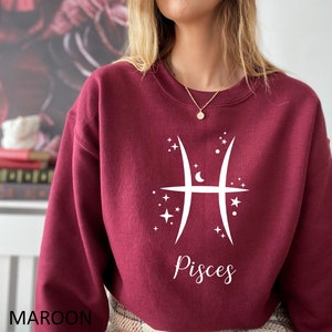 Op de afbeelding: Een bordeauxrood sweatshirt met een wit Vissen-sterrenbeeld en het woord "Vissen" gedrukt op de voorkant.
