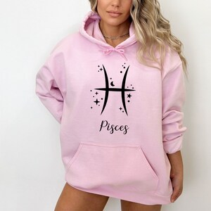 Op de afbeelding: Een roze hoodie met een zwart Vissen-sterrenbeeld en sterren. De tekst "Vissen" staat onder het symbool.