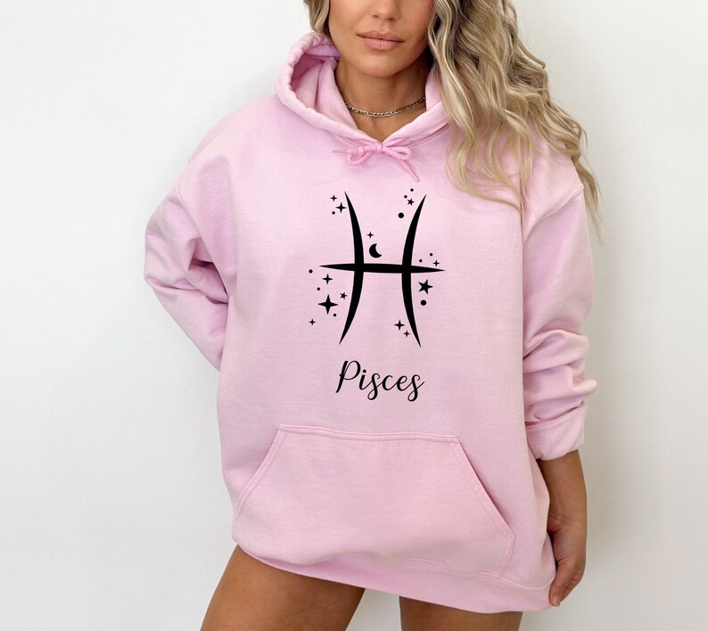 Op de afbeelding: Een roze hoodie met een zwart Vissen-sterrenbeeld en sterren. De tekst "Vissen" staat onder het symbool.