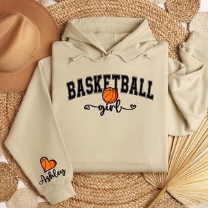 May include: Beige hoodie with a basketball graphic and the text "Basketball girl" on the front. The left sleeve has a heart graphic and the text "Ashley" on it.