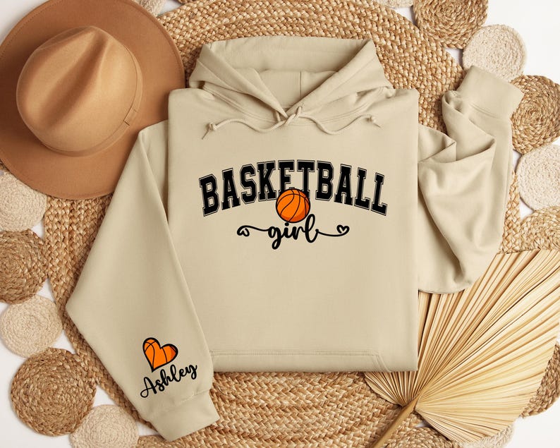 May include: Beige hoodie with a basketball graphic and the text "Basketball girl" on the front. The left sleeve has a heart graphic and the text "Ashley" on it.