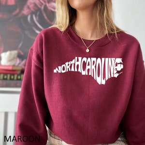 K&ouml;nnte beinhalten: Maroonfarbener Pullover mit einem wei&szlig;en Umriss des Bundesstaates North Carolina und dem Text "NORTH CAROLINA" innerhalb des Umrisses.