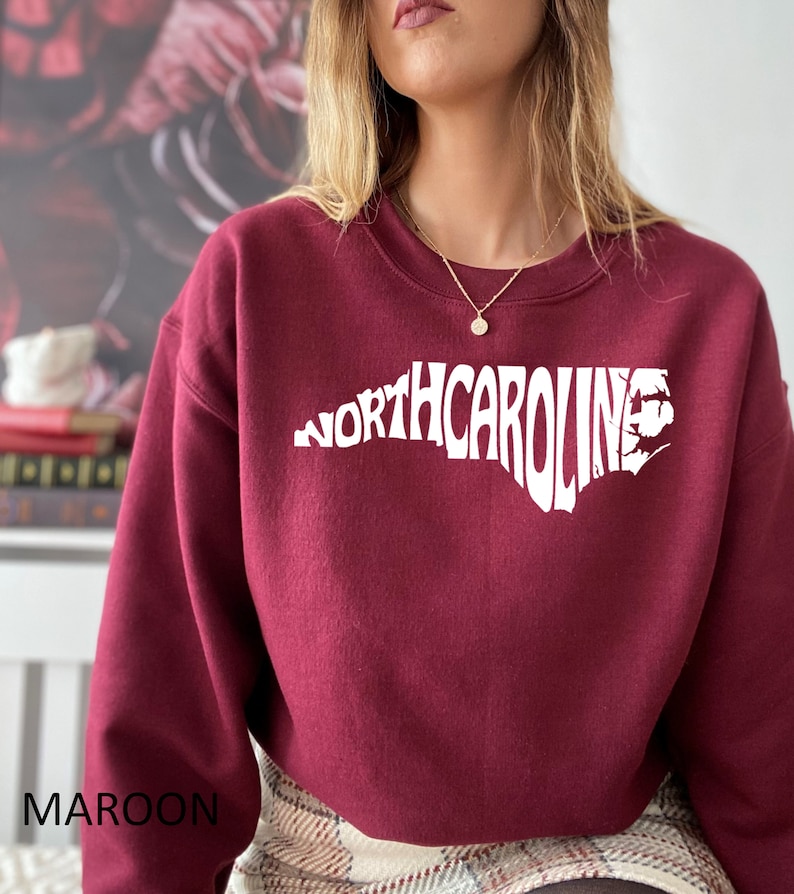 K&ouml;nnte beinhalten: Maroonfarbener Pullover mit einem wei&szlig;en Umriss des Bundesstaates North Carolina und dem Text "NORTH CAROLINA" innerhalb des Umrisses.