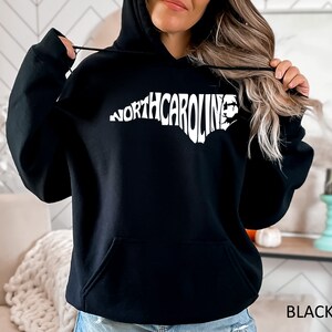 K&ouml;nnte beinhalten: Schwarzer Kapuzenpullover mit einem wei&szlig;en Umriss des Bundesstaates North Carolina und dem Text "NORTH CAROLINA" innerhalb des Umrisses.