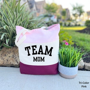 May include: A white canvas tote bag with pink and burgundy accents. The bag has a black graphic that says "TEAM MOM".