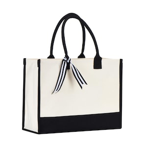 Puede incluir: Una bolsa de lona blanca con ribetes negros y asas negras. La bolsa tiene una cinta de rayas blancas y negras atada alrededor de las asas.