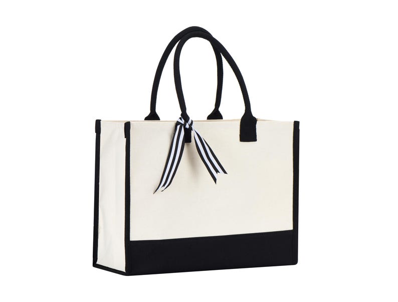 Puede incluir: Una bolsa de lona blanca con ribetes negros y asas negras. La bolsa tiene una cinta de rayas blancas y negras atada alrededor de las asas.