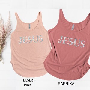 May include: Two tank tops with the word "JESUS" spelled out in floral letters. The tank top on the left is a light pink color and the tank top on the right is a reddish-brown color.