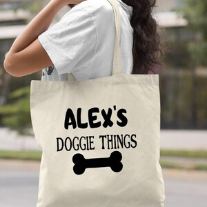 A woman carrying a white tote bag with the text "ALEX'S DOGGIE THINGS" printed on it.