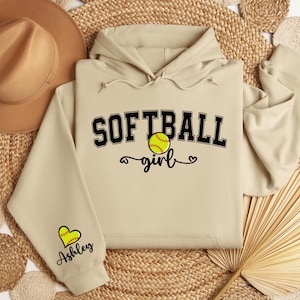 May include: A beige hoodie with the text "Softball girl" and a softball graphic. The hoodie has a heart with the name "Ashley" on the sleeve.