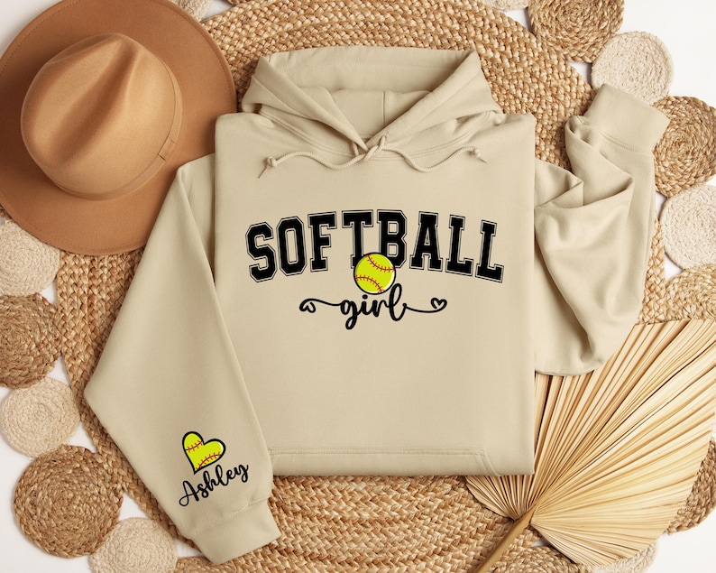 May include: A beige hoodie with the text "Softball girl" and a softball graphic. The hoodie has a heart with the name "Ashley" on the sleeve.