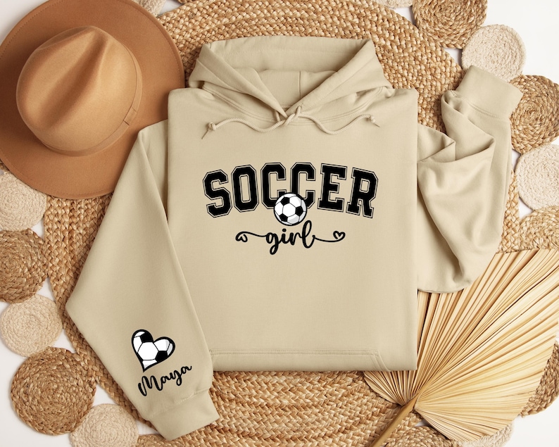 May include: A beige hoodie with the text "SOCCER girl" and a soccer ball graphic on the front. The left sleeve has a heart with a soccer ball graphic and the name "Maya" underneath.