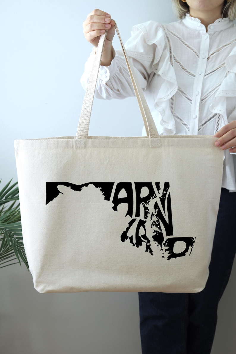 May include: A cream-colored tote bag with black graphic text and a map outline of Maryland. The bag has long, sturdy straps for carrying and is made of a durable fabric.