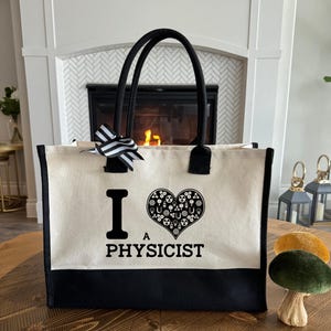 May include: A canvas tote bag with black handles and a black bottom. The bag features the text "I A PHYSICIST" with a heart-shaped graphic filled with physics-related symbols. A black and white striped bow is attached to the top.