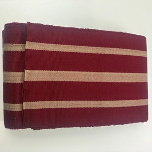 Può includere: Rotolo di tessuto bordeaux a righe orizzontali beige. Il tessuto è a trama fitta, potenzialmente adatto per tappezzeria o arredamento. Le strisce sono uniformemente distanziate.