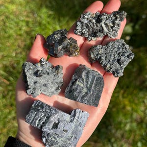 May include: A collection of six black and silver crystals, some with a green hue, on a hand. The crystals are various shapes and sizes.