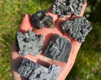 May include: A collection of six black and silver crystals, some with a green hue, on a hand. The crystals are various shapes and sizes.
