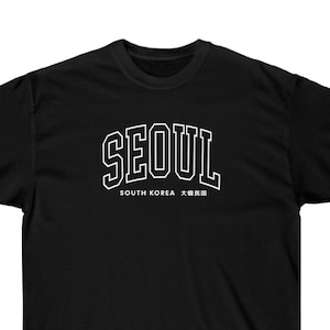 May include: Black t-shirt with white text that reads "SEOUL" in a bold, collegiate-style font. Below the text is "SOUTH KOREA" and the Korean characters for "Seoul".