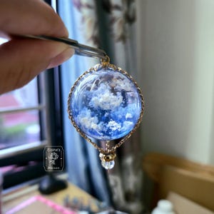 Puede incluir: Un colgante artesanal con una esfera de resina transparente con un diseño de cielo azul y nubes blancas esponjosas. La esfera está encerrada en un marco y cadena de metal dorado. El colgante está sujeto con unas pinzas.