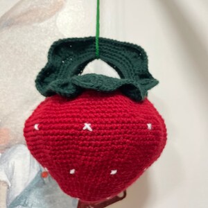 Könnte beinhalten: Ein gehäkeltes, hängendes Ornament in Form einer Erdbeere mit einem grünen, gehäkelten Blatt oben. Die Erdbeere ist rot mit weißen Stichen für die Samen.