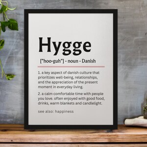 May include: A black framed print with a white background and the word "Hygge" in bold black text. The definition of the word is in smaller black text, with the pronunciation, "[hoo-guh]", and the word's origin, "Danish". The definition is: "A key aspect of Danish culture that prioritizes well-being, relationships, and the appreciation of the present moment in everyday living. A calm comfortable time with people you love. Often enjoyed with good food, drinks, warm blankets and candlelight. See also: happiness."
